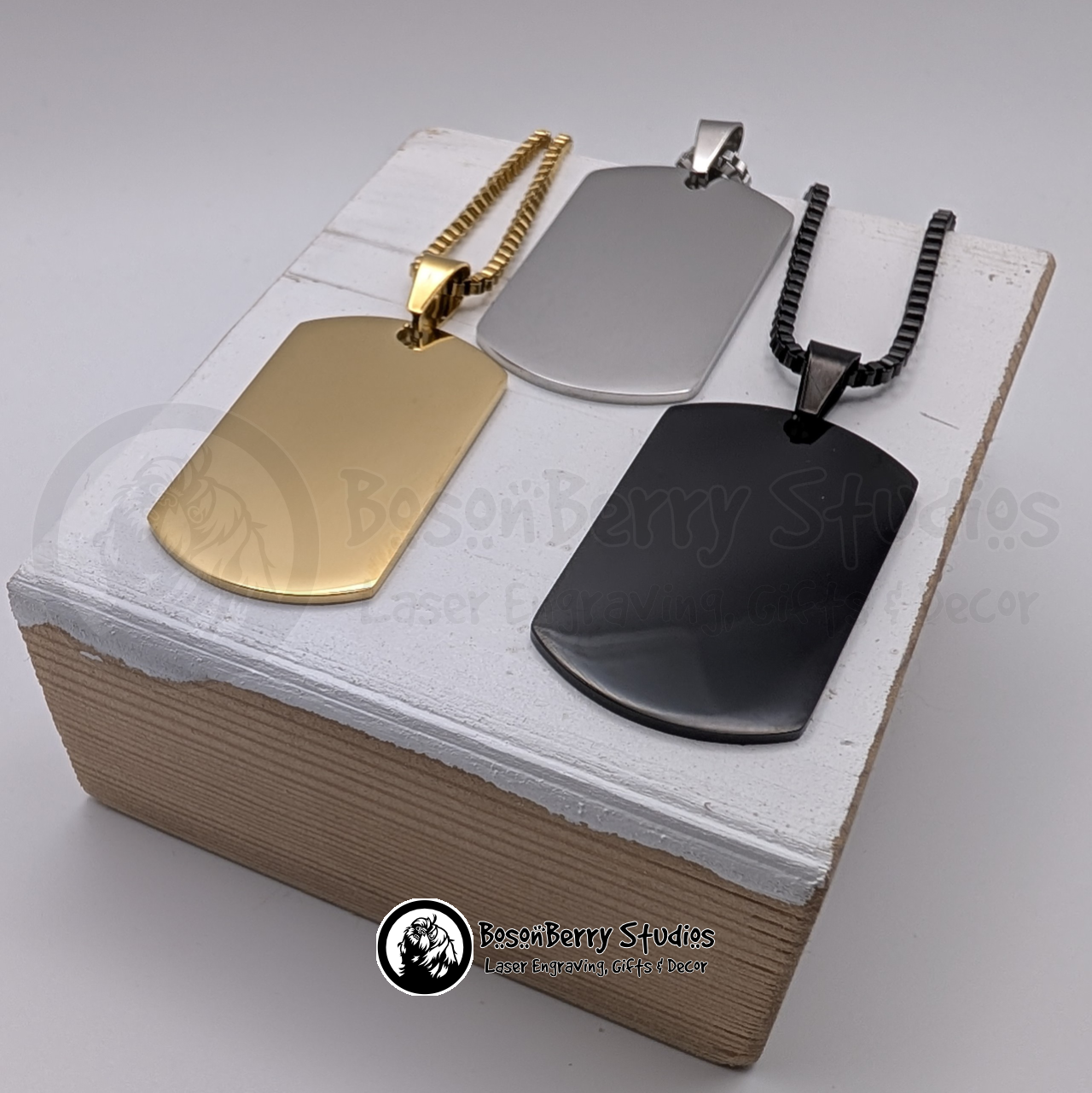 Three dog tags in gold, silver, and black on a white wooden stand with 'BosonBerry Studios' branding.