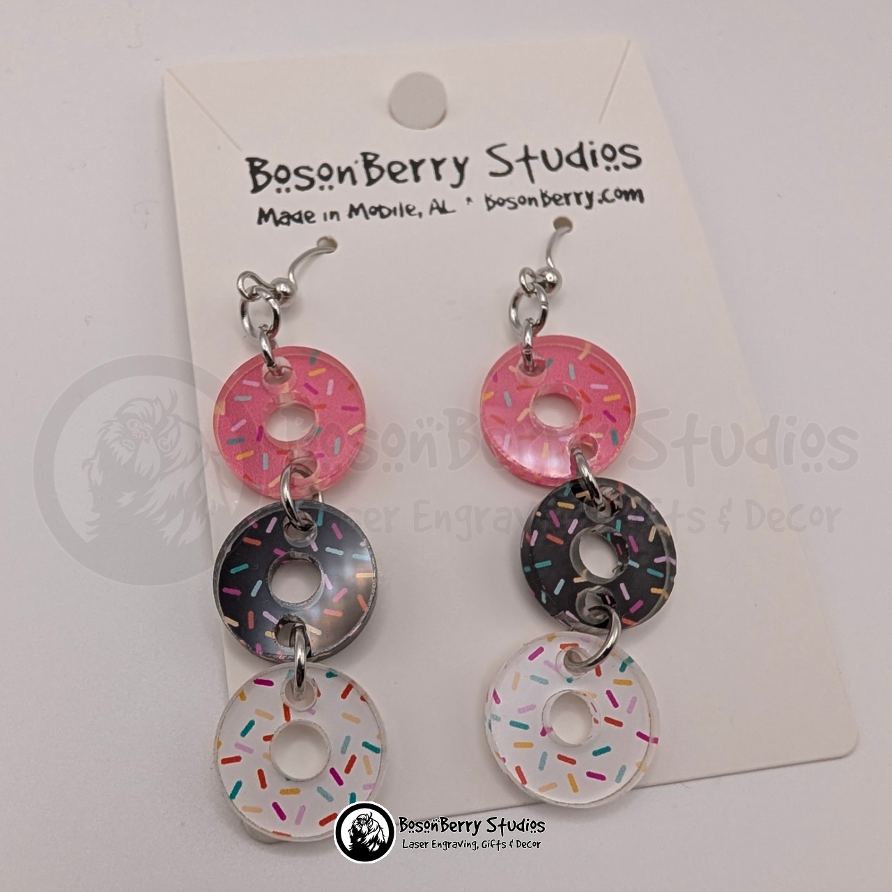 A trio of pink, white, and chocolate sprinkle donut-shaped acrylic pieces dangle from each earring on a BosonBerry Studios branded card.