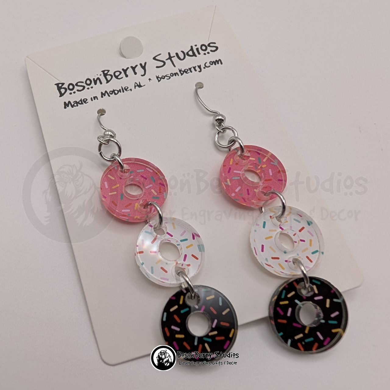 A trio of pink, white, and chocolate sprinkle donut-shaped acrylic pieces dangle from each earring on a BosonBerry Studios branded card.
