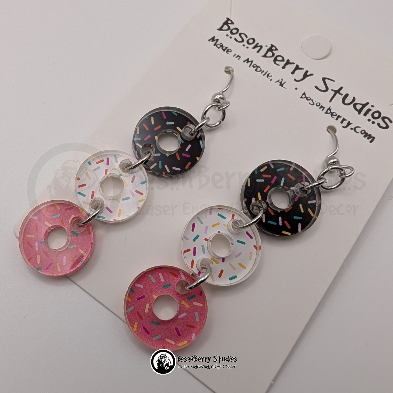 A trio of pink, white, and chocolate sprinkle donut-shaped acrylic pieces dangle from each earring on a BosonBerry Studios branded card.