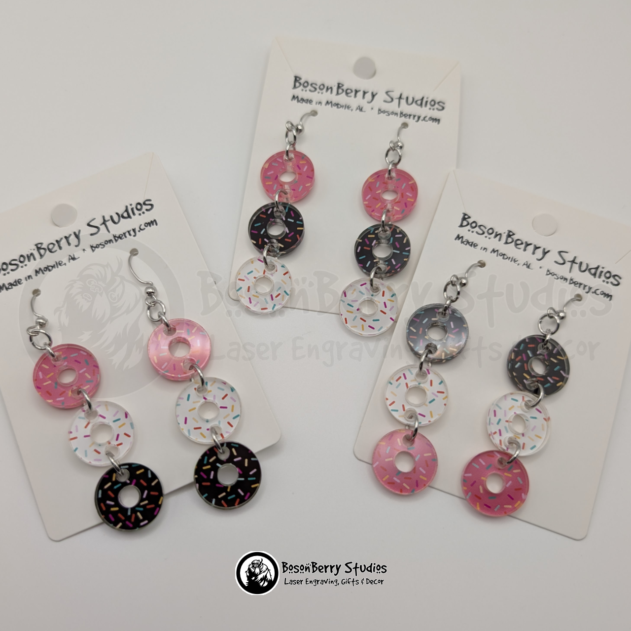 A trio of pink, white, and chocolate sprinkle donut-shaped acrylic pieces dangle from each earring on a BosonBerry Studios branded card.