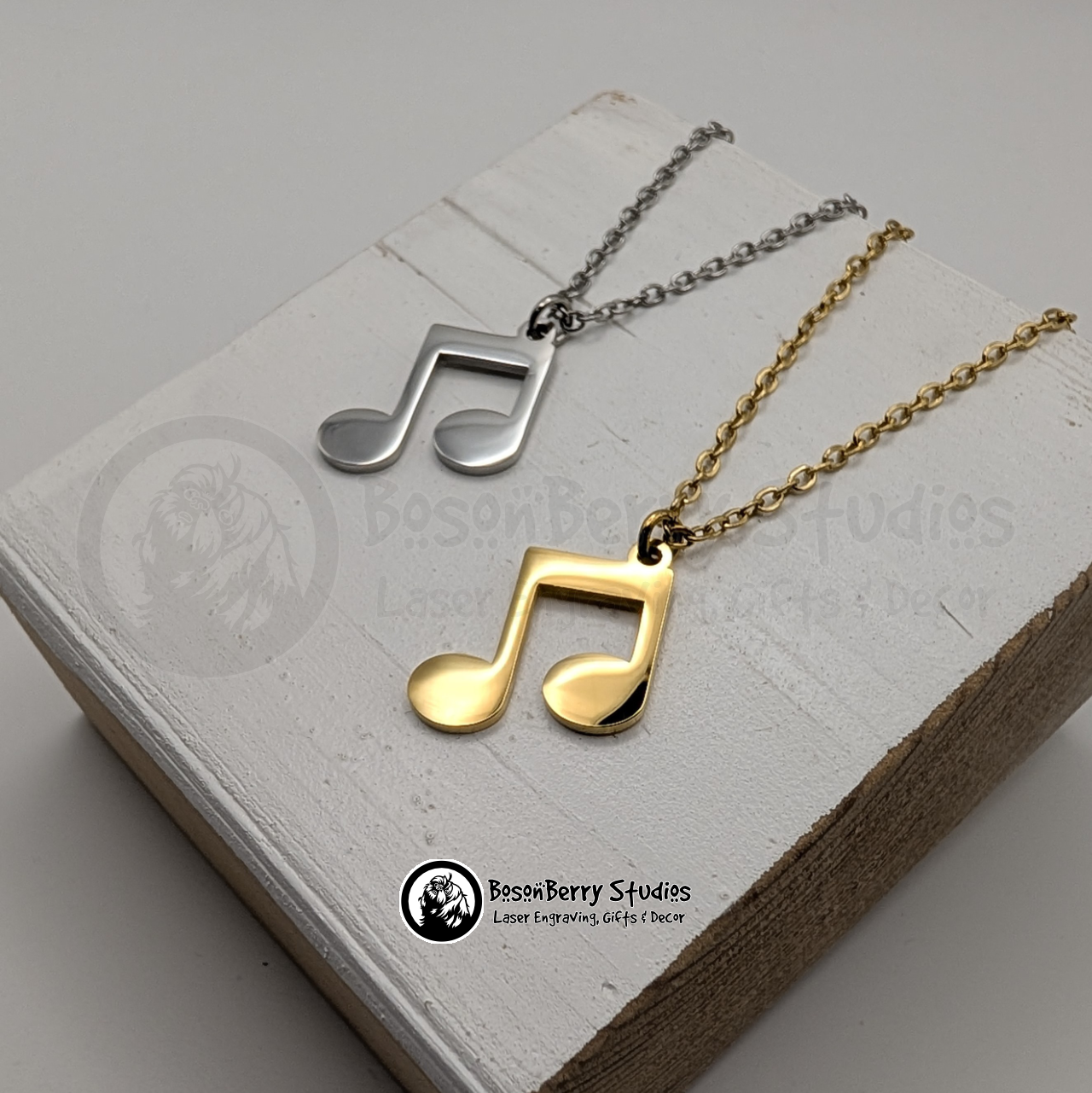 Two necklaces with music note pendants, one gold and one silver, on a white wooden stand with 'BosonBerry Studios' branding.