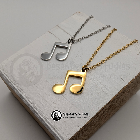 Two necklaces with music note pendants, one gold and one silver, on a white wooden stand with 'BosonBerry Studios' branding.