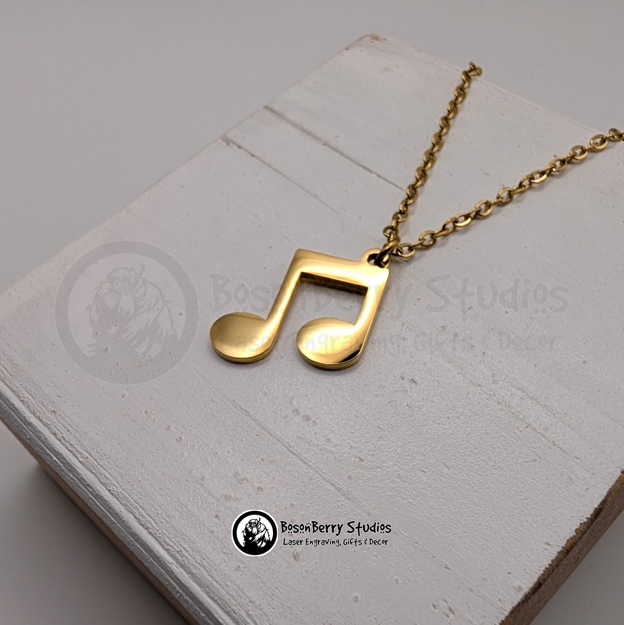 Gold music note pendant necklace on a white wooden stand with 'BosonBerry Studios' branding.