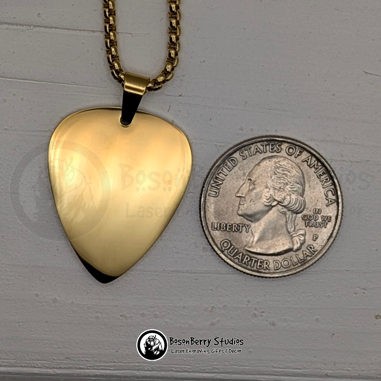 Gold guitar pick-shaped pendant necklace, next to a quarter for scale, on a white wooden stand with 'BosonBerry Studios' branding.