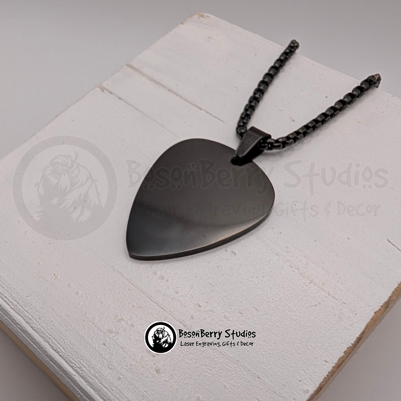 Black guitar pick-shaped pendant necklace on a white wooden stand with 'BosonBerry Studios' branding.