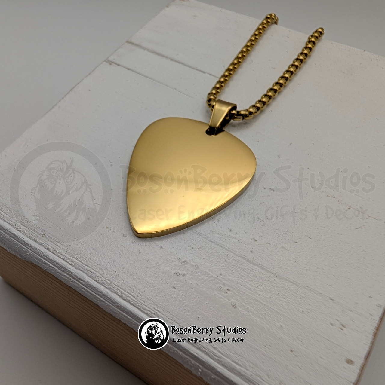 Gold guitar pick-shaped pendant necklace on a white wooden stand with 'BosonBerry Studios' branding.