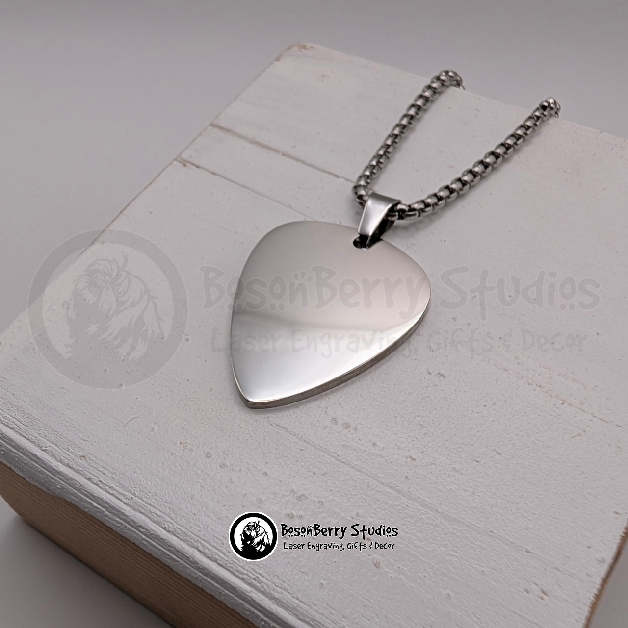 Silver guitar pick-shaped pendant necklace on a white wooden stand with 'BosonBerry Studios' branding.