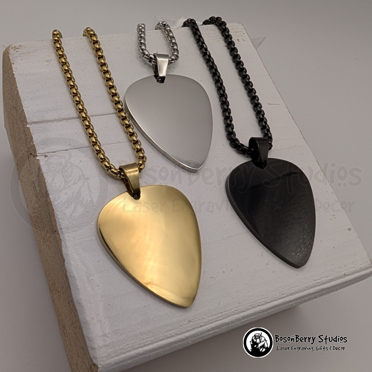 Three necklaces, gold, silver and black, with guitar pick-shaped pendants on a white wooden stand with 'BosonBerry Studios' branding.