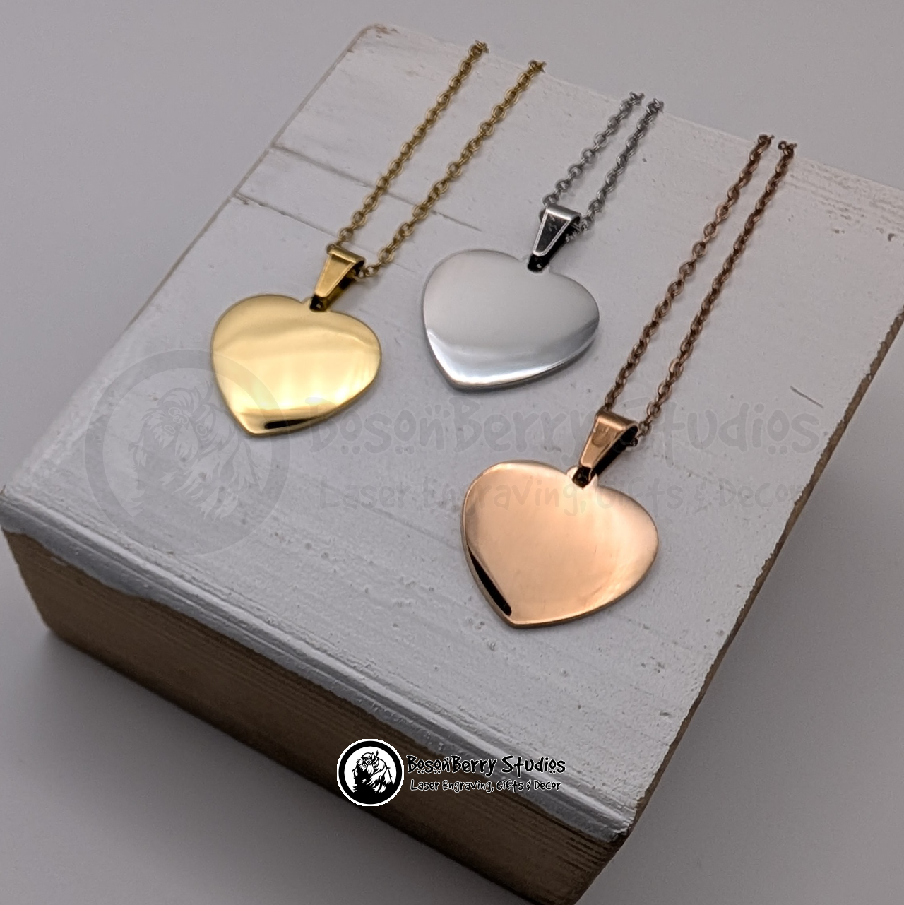 Trio of heart-shaped pendant necklaces, gold, silver, and rose gold, on a white wooden stand with 'BosonBerry Studios' branding.