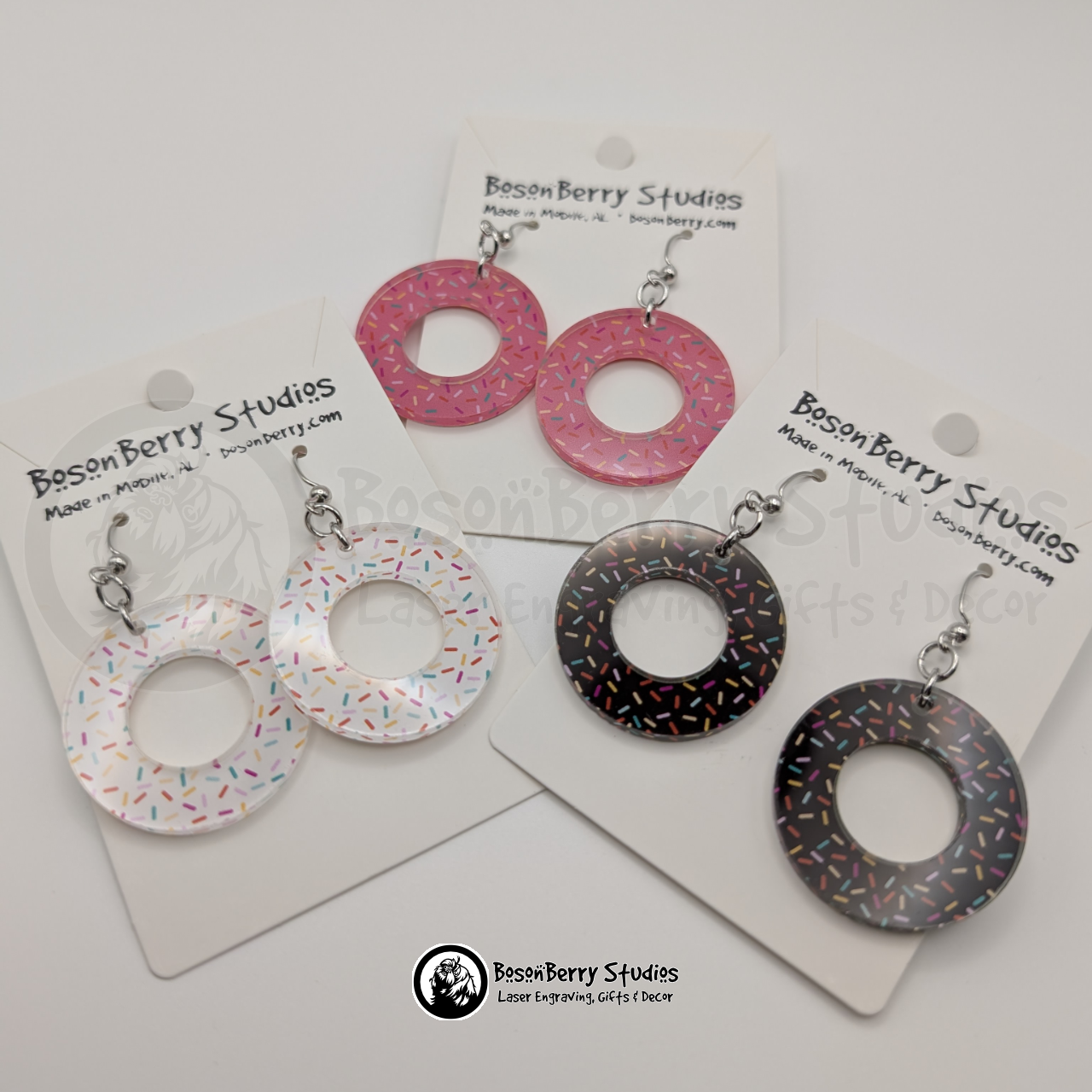 Trio of large chocolate donut-shaped acrylic earrings, one chocolate, one white and one pink, with a colorful sprinkle pattern, on a BosonBerry Studios card.