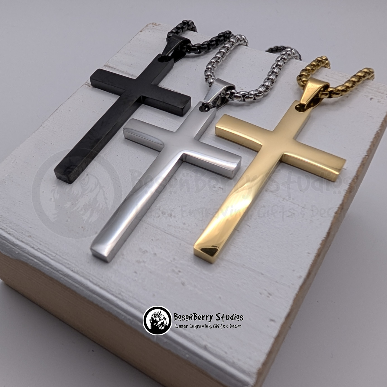 Three large cross pendants in black, silver, and gold on a white wooden stand with 'BosonBerry Studios' branding.