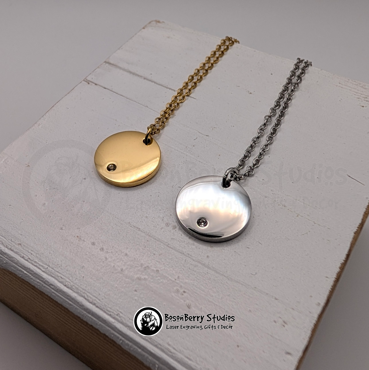 One gold and one silver necklace with round pendants inset with tiny cubic zirconia crystals on a white wooden stand with 'BosonBerry Studios' branding.