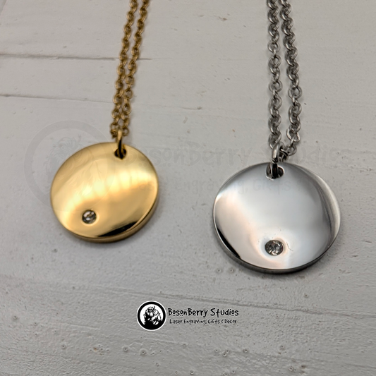 Two necklaces with round pendants with a tiny inset cubic zirconia crystal, one gold and one silver, on a white wooden stand with 'BosonBerry Studios' branding.