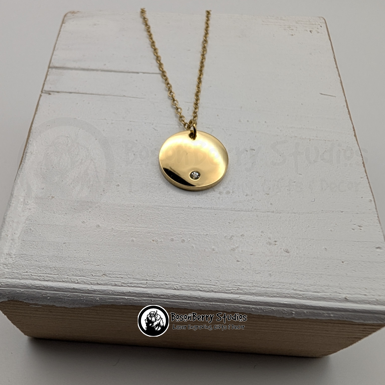 Gold necklace with a round pendant inset with a tiny cubic zirconia crystal on a white wooden stand with 'BosonBerry Studios' branding.