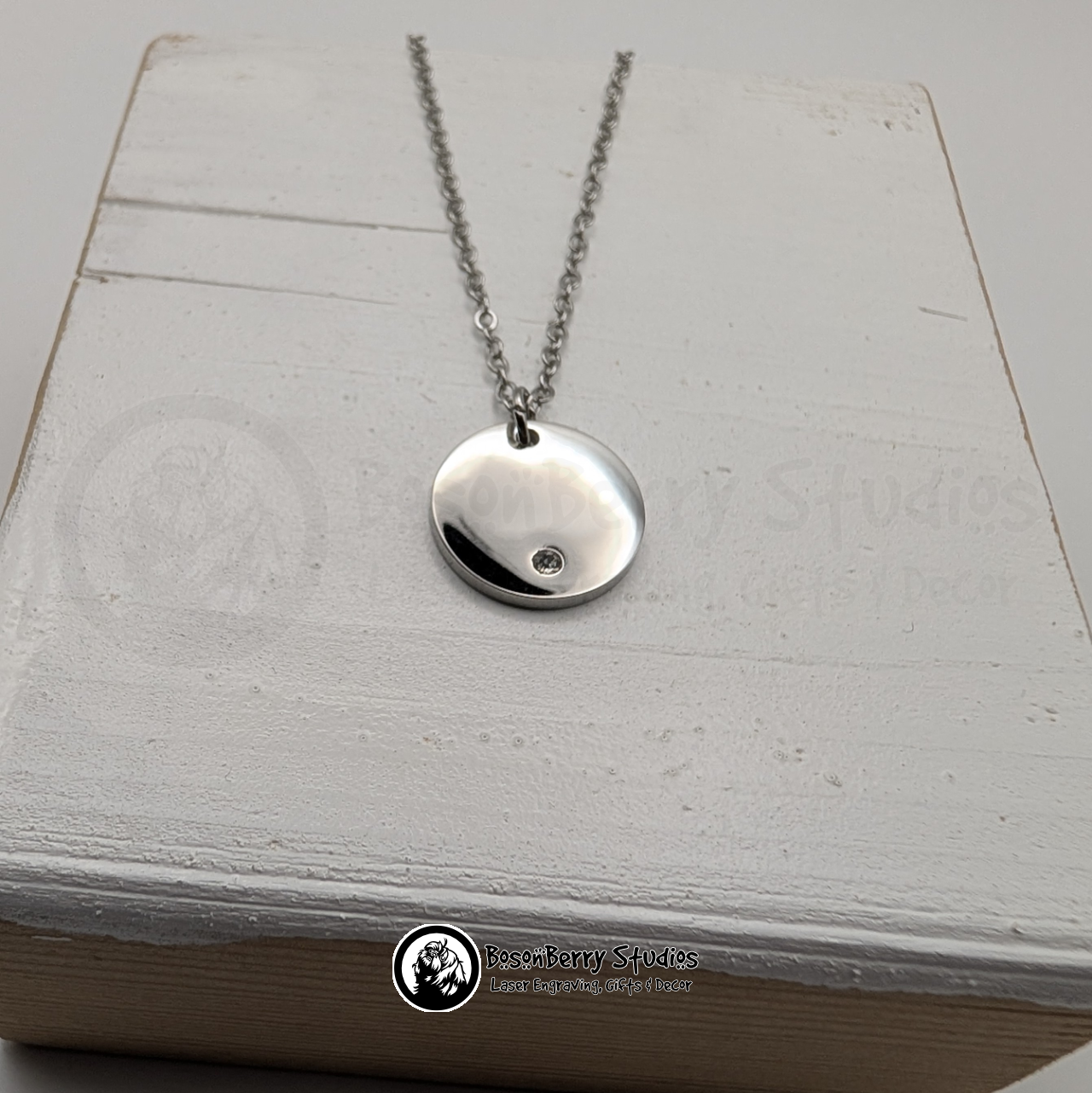 Silver necklace with a round pendant inset with a tiny cubic zirconia crystal on a white wooden stand with 'BosonBerry Studios' branding.