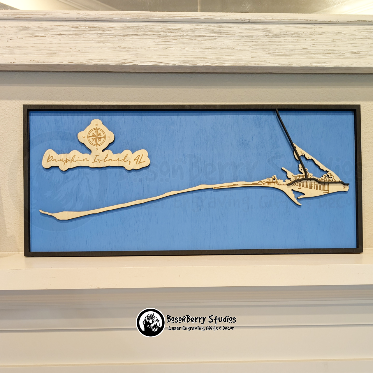 Wooden layered map of Dauphin Island, Alabama with a black frame and major roads, wooden land engraved with minor roads, and blue water, with 'BosonBerry Studios' branding.