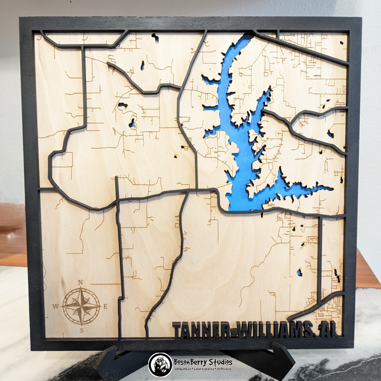 Wooden layered map of Tanner Williams, Alabama with a black frame and major roads, wooden land engraved with minor roads, and blue water, with 'BosonBerry Studios' branding.