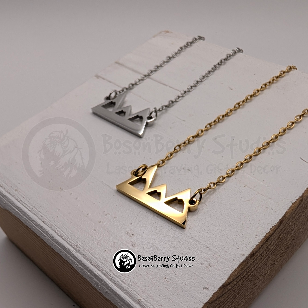 Two necklaces with mountain pendants, one gold and one silver, on a white wooden stand with 'BosonBerry Studios' branding.
