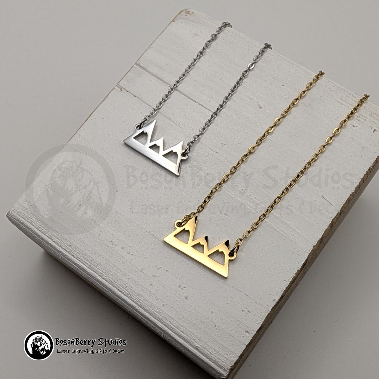 Two necklaces with mountain pendants, one gold and one silver, on a white wooden stand with 'BosonBerry Studios' branding.