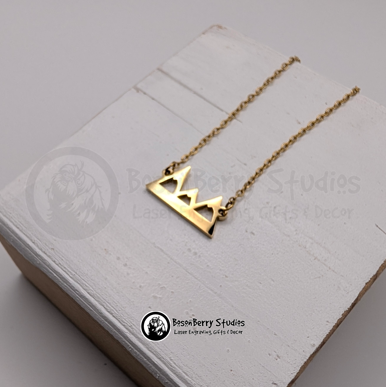 Gold necklace with a mountain pendant on a white wooden stand with 'BosonBerry Studios' branding.