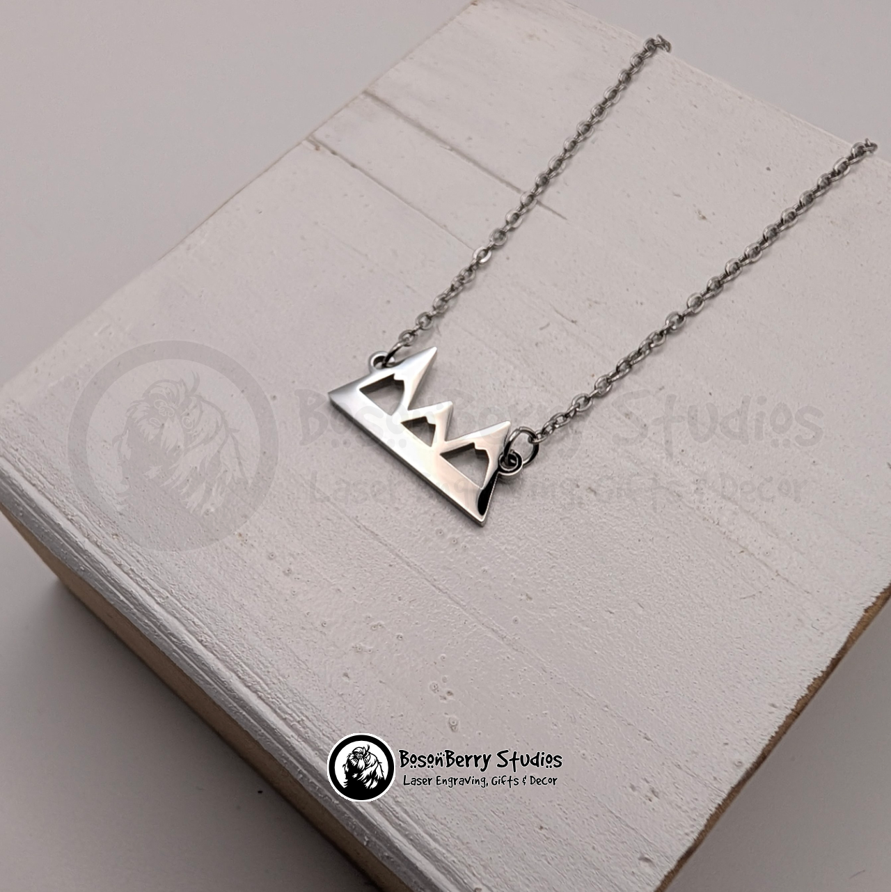 Silver necklace with a mountain pendant on a white wooden stand with 'BosonBerry Studios' branding.