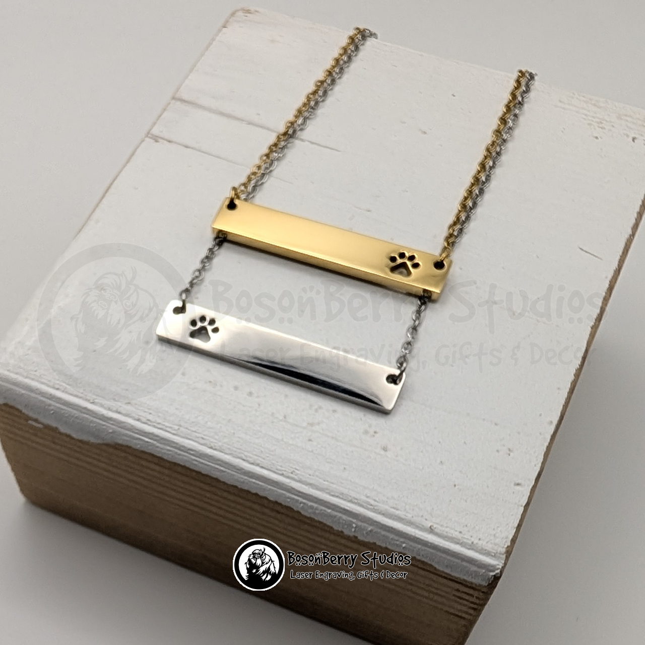 Two necklaces with bar pendants featuring paw print-shaped cutouts, one gold and one silver, on a white wooden stand with 'BosonBerry Studios' branding.