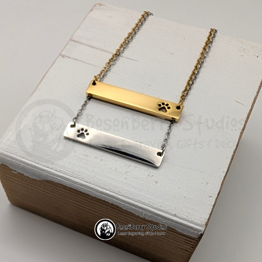Two necklaces with bar pendants featuring paw print-shaped cutouts, one gold and one silver, on a white wooden stand with 'BosonBerry Studios' branding.