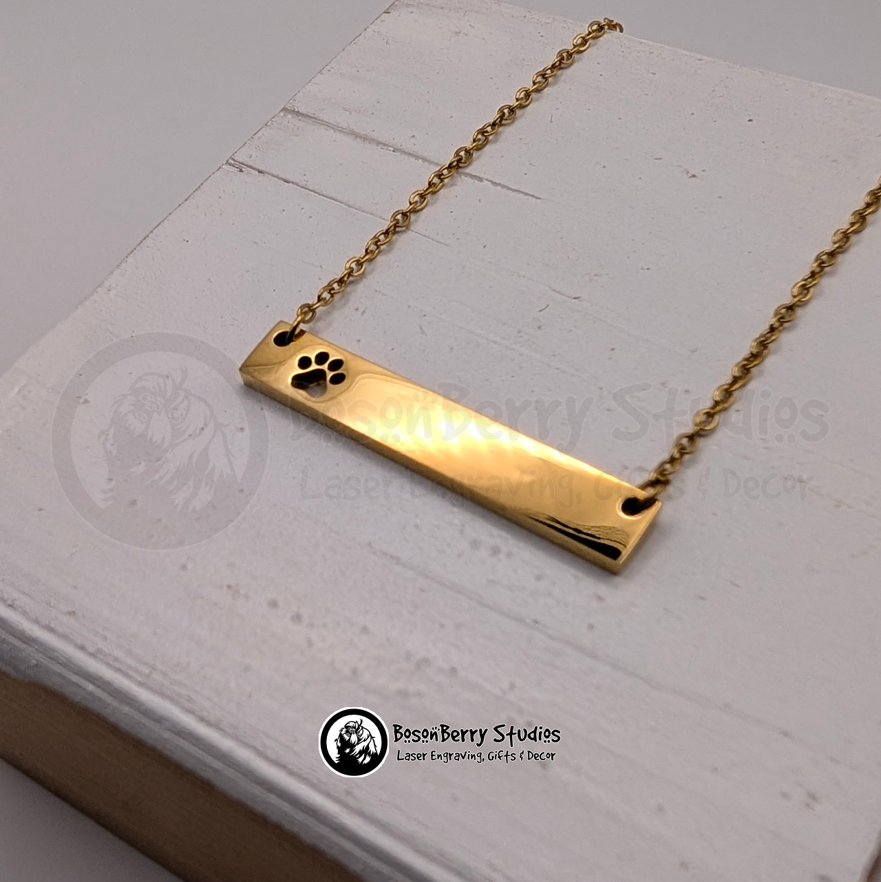 Gold necklace with bar pendant featuring paw print-shaped cutout on a white wooden stand with 'BosonBerry Studios' branding.