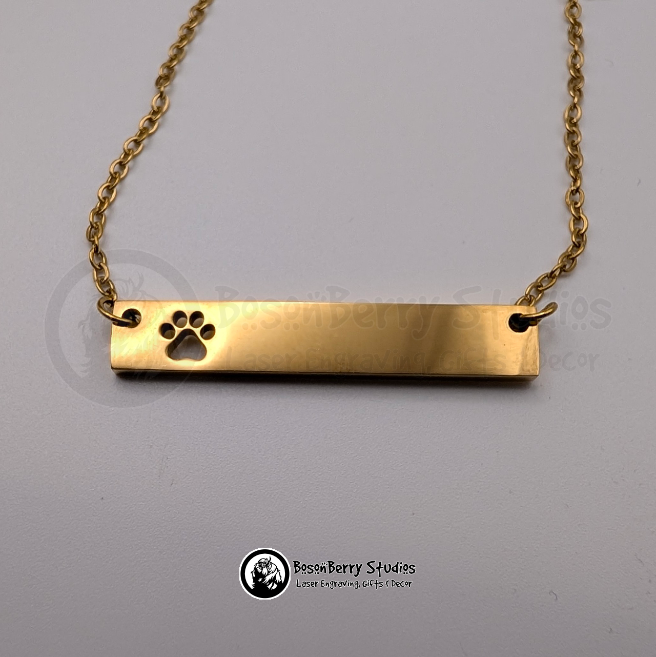 Gold necklace with bar pendant featuring paw print-shaped cutout on a white background with 'BosonBerry Studios' branding.