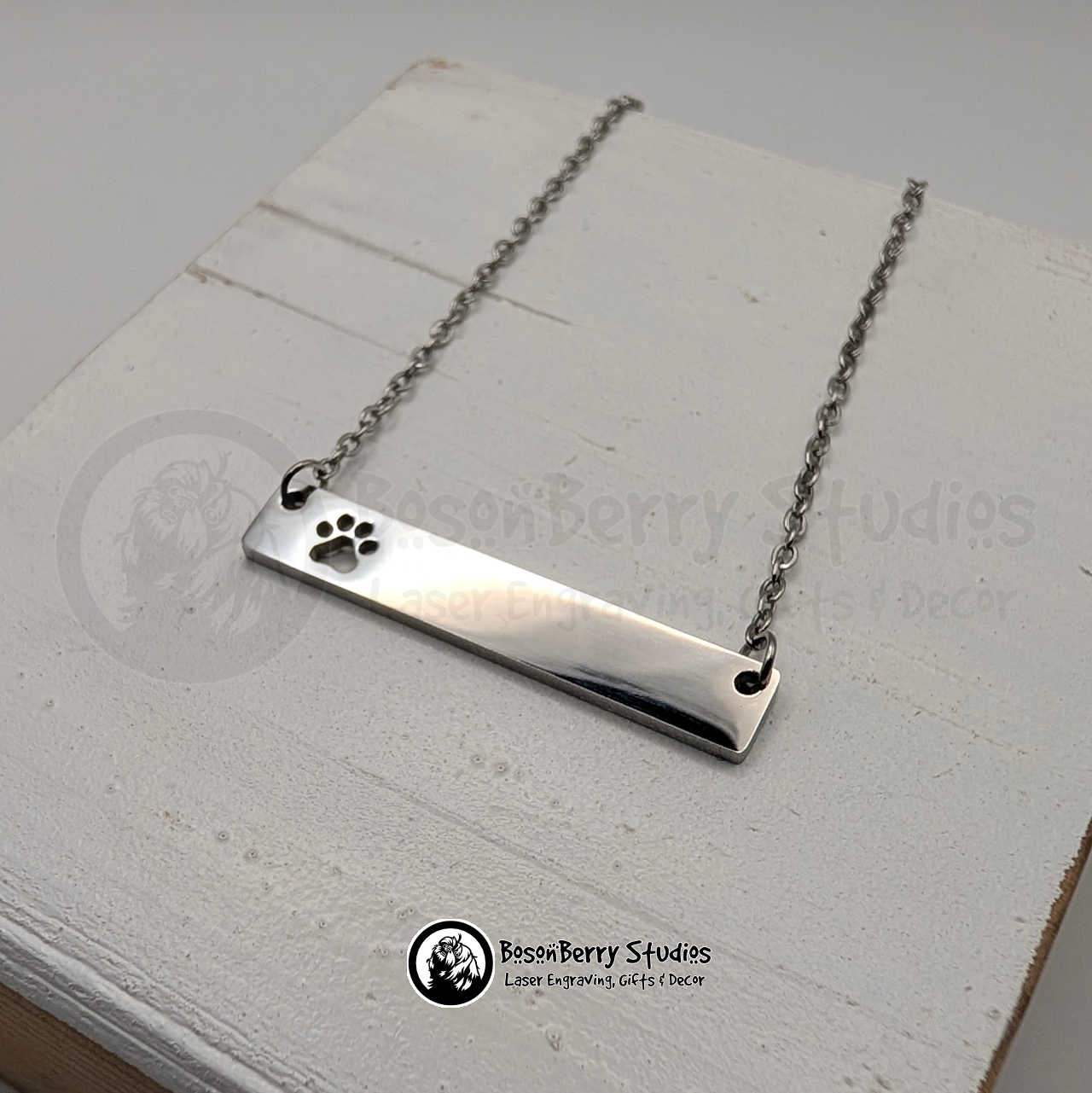 Silver necklace with bar pendant featuring paw print-shaped cutout on a white wooden stand with 'BosonBerry Studios' branding.