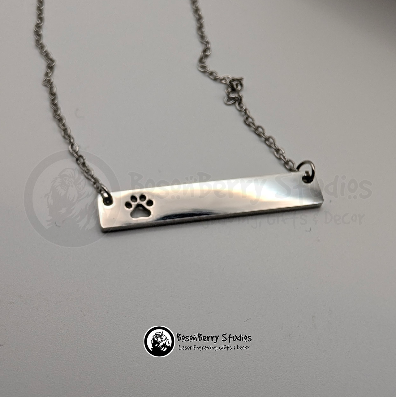 Silver necklace with bar pendant featuring paw print-shaped cutout on a white background with 'BosonBerry Studios' branding.