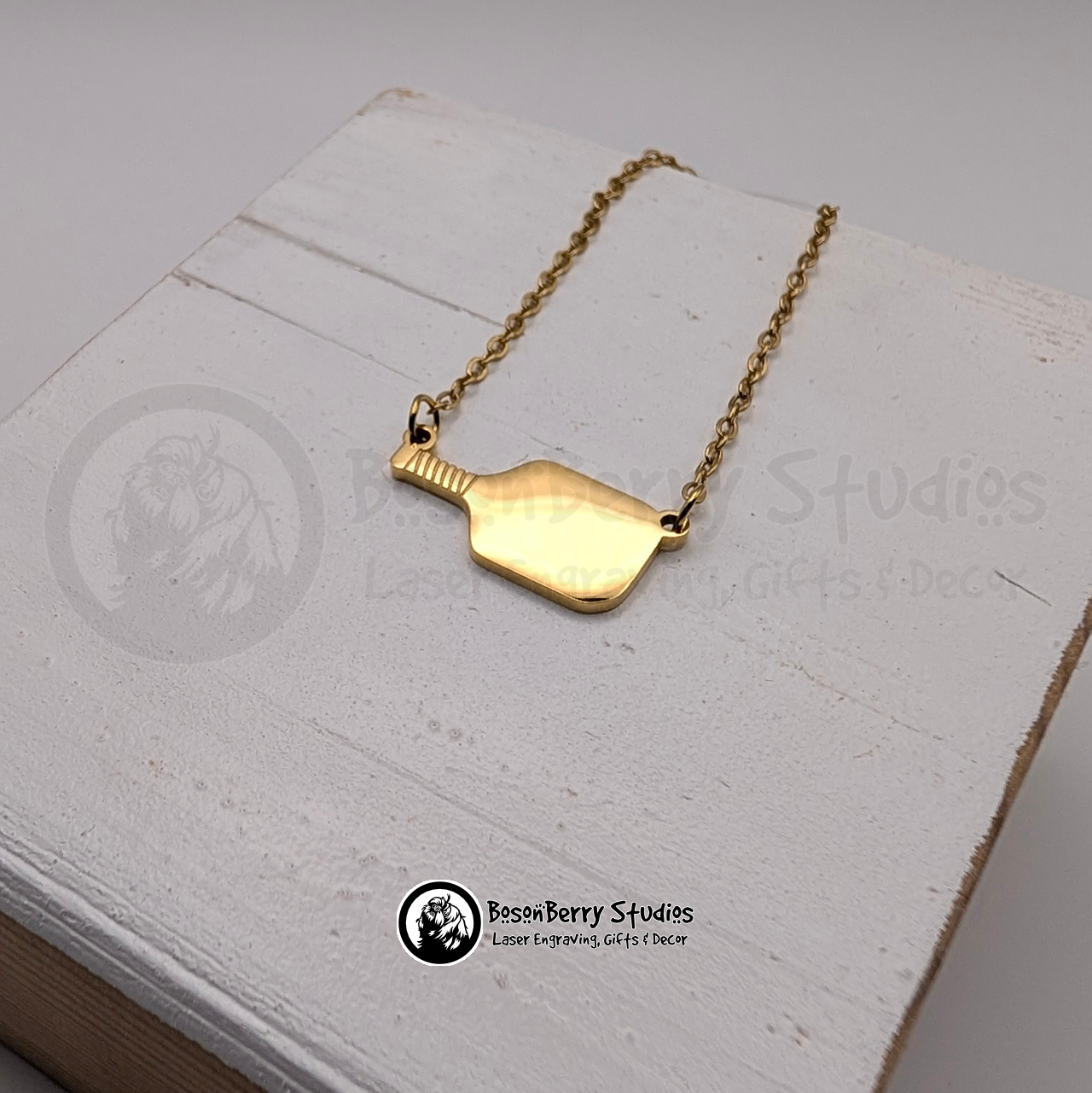 Gold pickleball paddle necklace on a white wooden stand with 'BosonBerry Studios' branding.