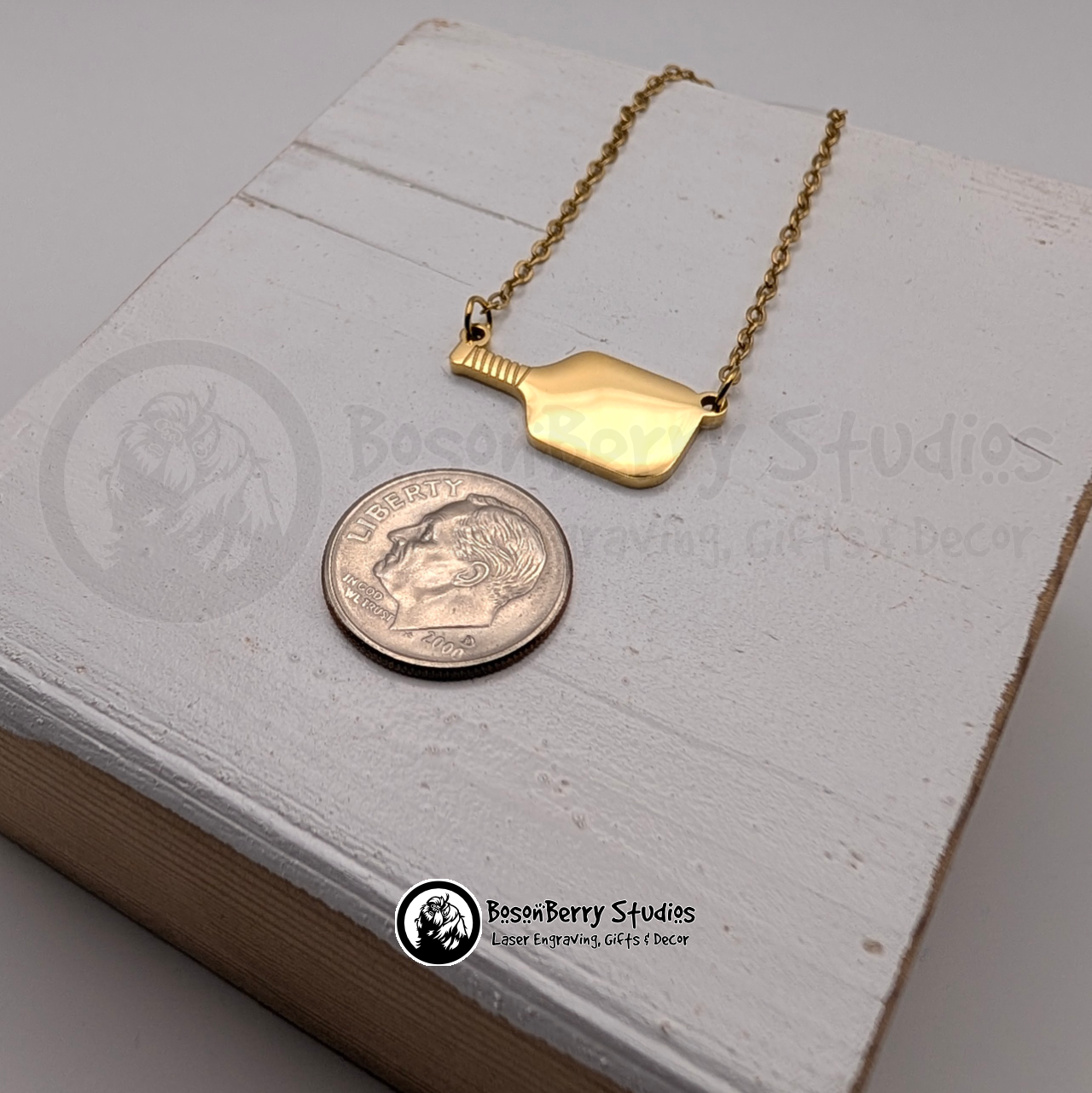 Gold pickleball paddle necklace, next to a dime for scale, on a white wooden stand with 'BosonBerry Studios' branding.