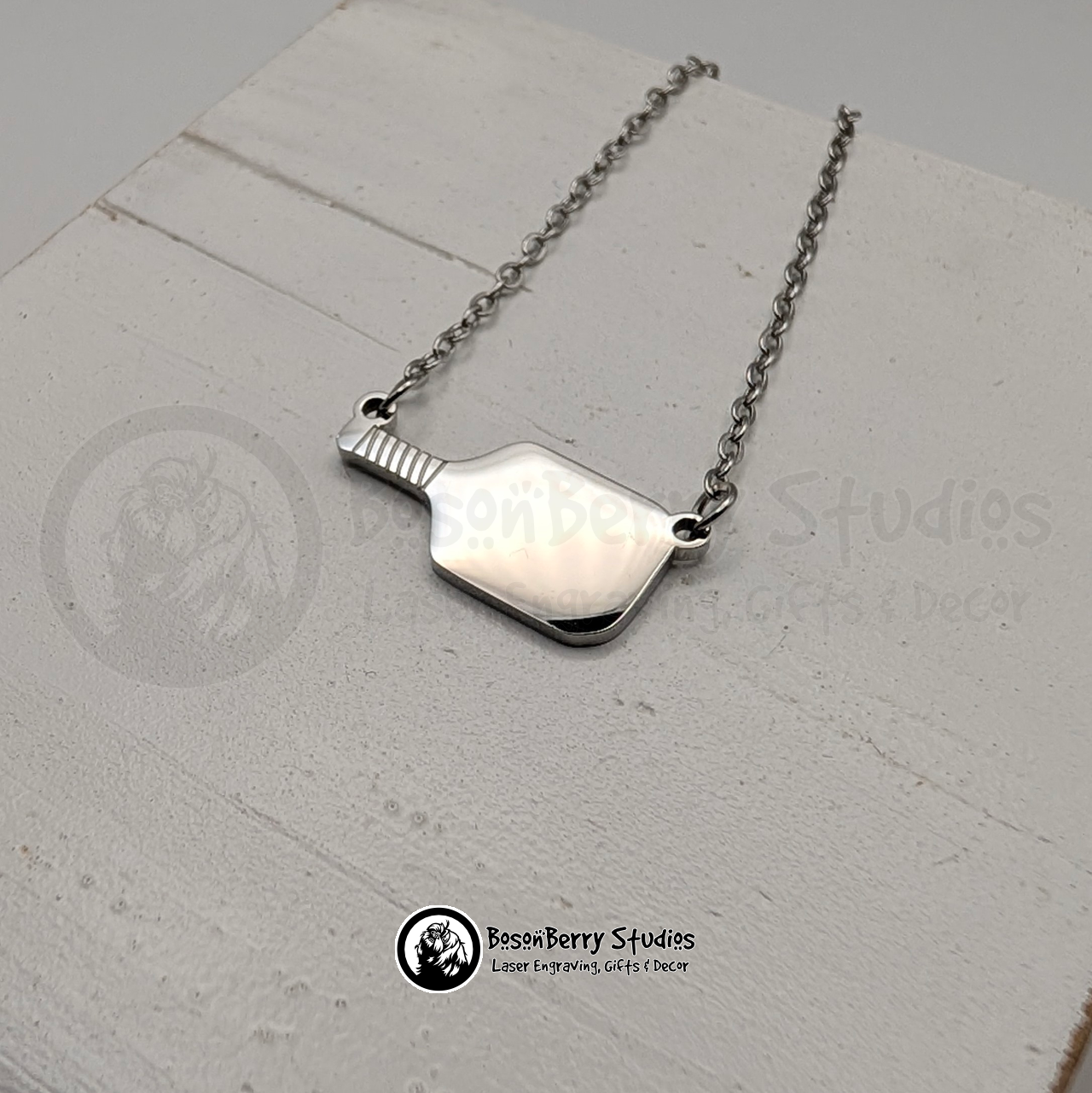 Silver pickleball paddle necklace on a white wooden stand with 'BosonBerry Studios' branding.