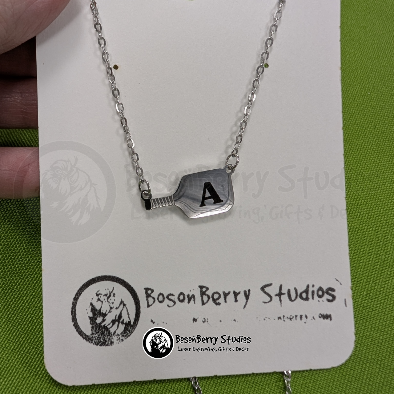 Silver pickleball paddle necklace engraved with a black letter 'A' on a white jewelry card with 'BosonBerry Studios' branding.