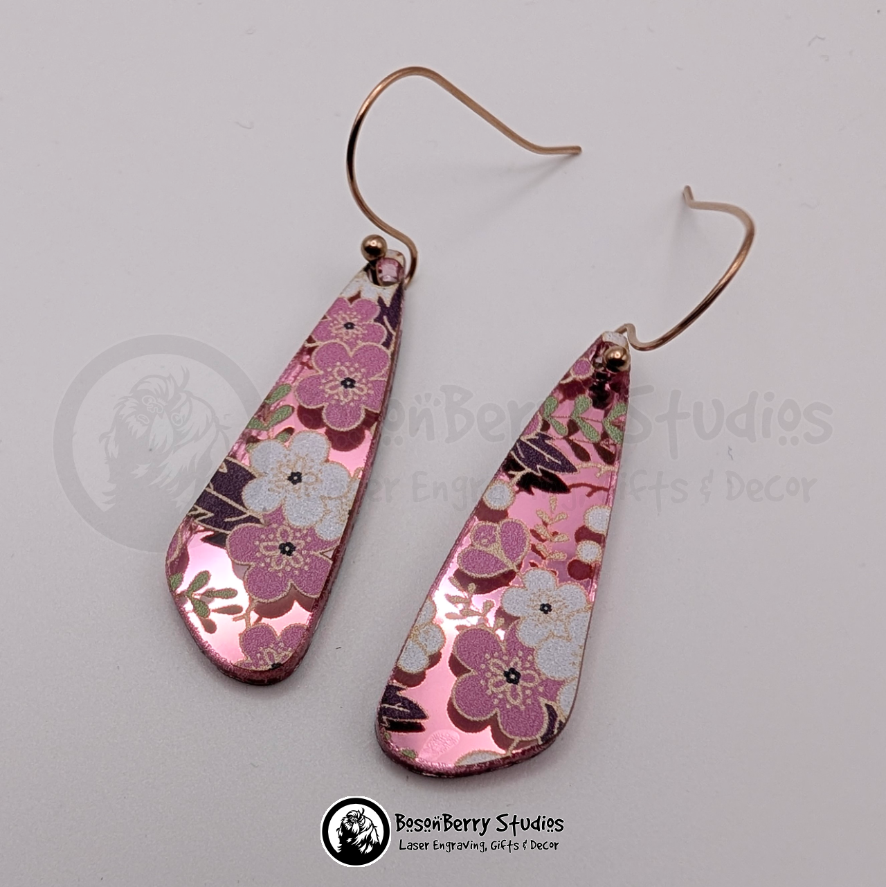 Earrings featuring a white and pink cherry blossom pattern on a pink mirrored acrylic field, in a skinny elongated teardrop shape, on a white background with 'BosonBerry Studios' branding.