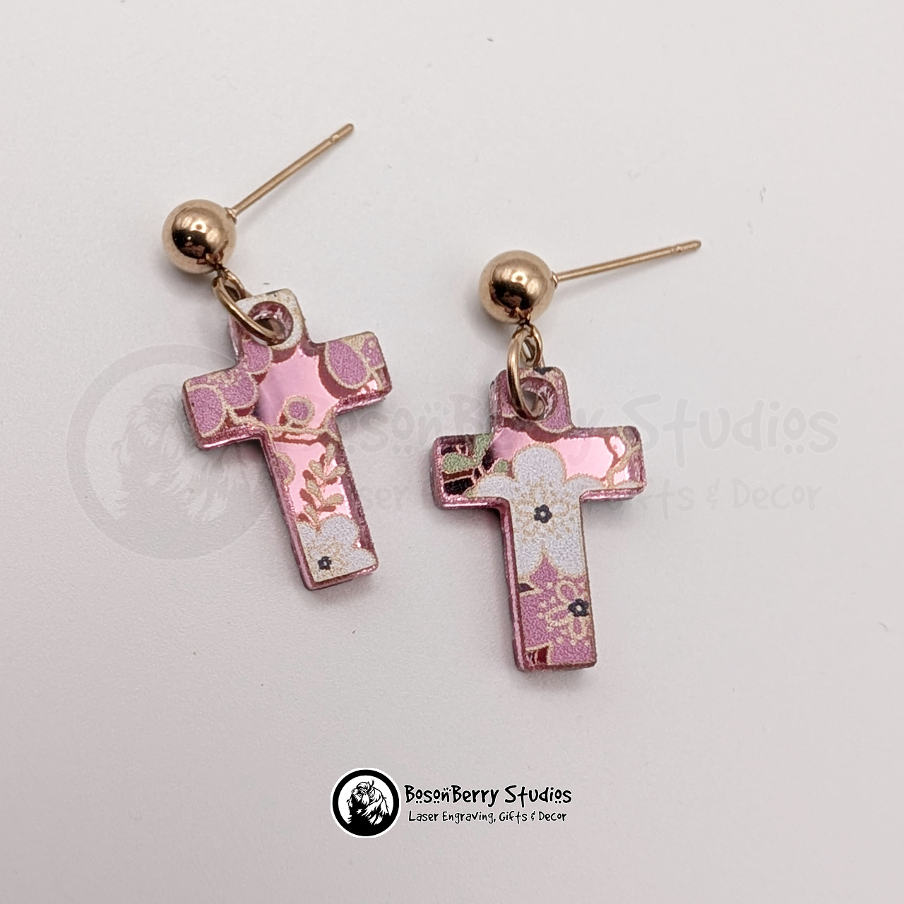 Earrings featuring a white and pink cherry blossom pattern on a pink mirrored acrylic field, in a tiny cross shape, on a white background with 'BosonBerry Studios' branding.