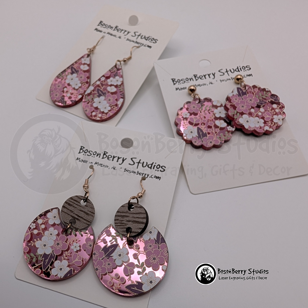Earrings featuring a white and pink cherry blossom pattern on a pink mirrored acrylic field, in teardrop, scalloped circle, and tiered disc shapes, on display cards with 'BosonBerry Studios' branding.