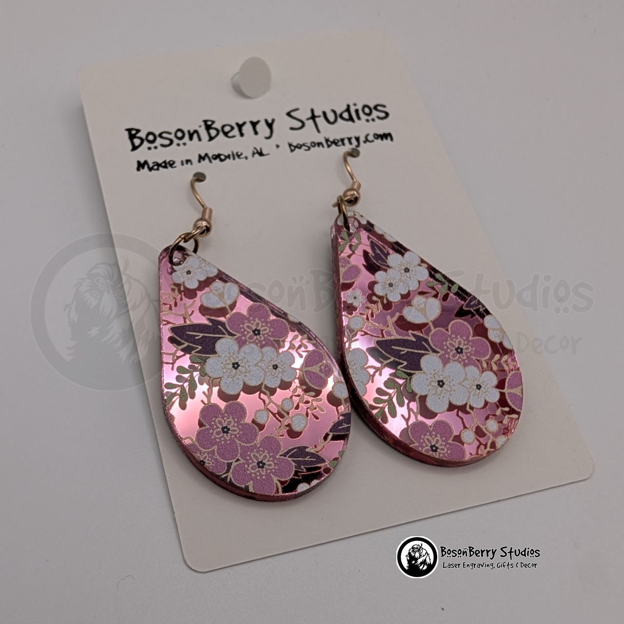 Earrings featuring a white and pink cherry blossom pattern on a pink mirrored acrylic field, in a teardrop shape, on display cards with 'BosonBerry Studios' branding.