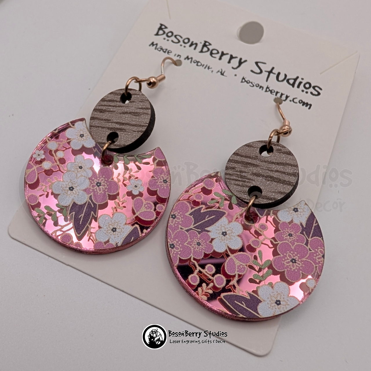 Earrings featuring a white and pink cherry blossom pattern on a pink mirrored acrylic field, in a disc shape suspended by a brown wooden connector, on display cards with 'BosonBerry Studios' branding.
