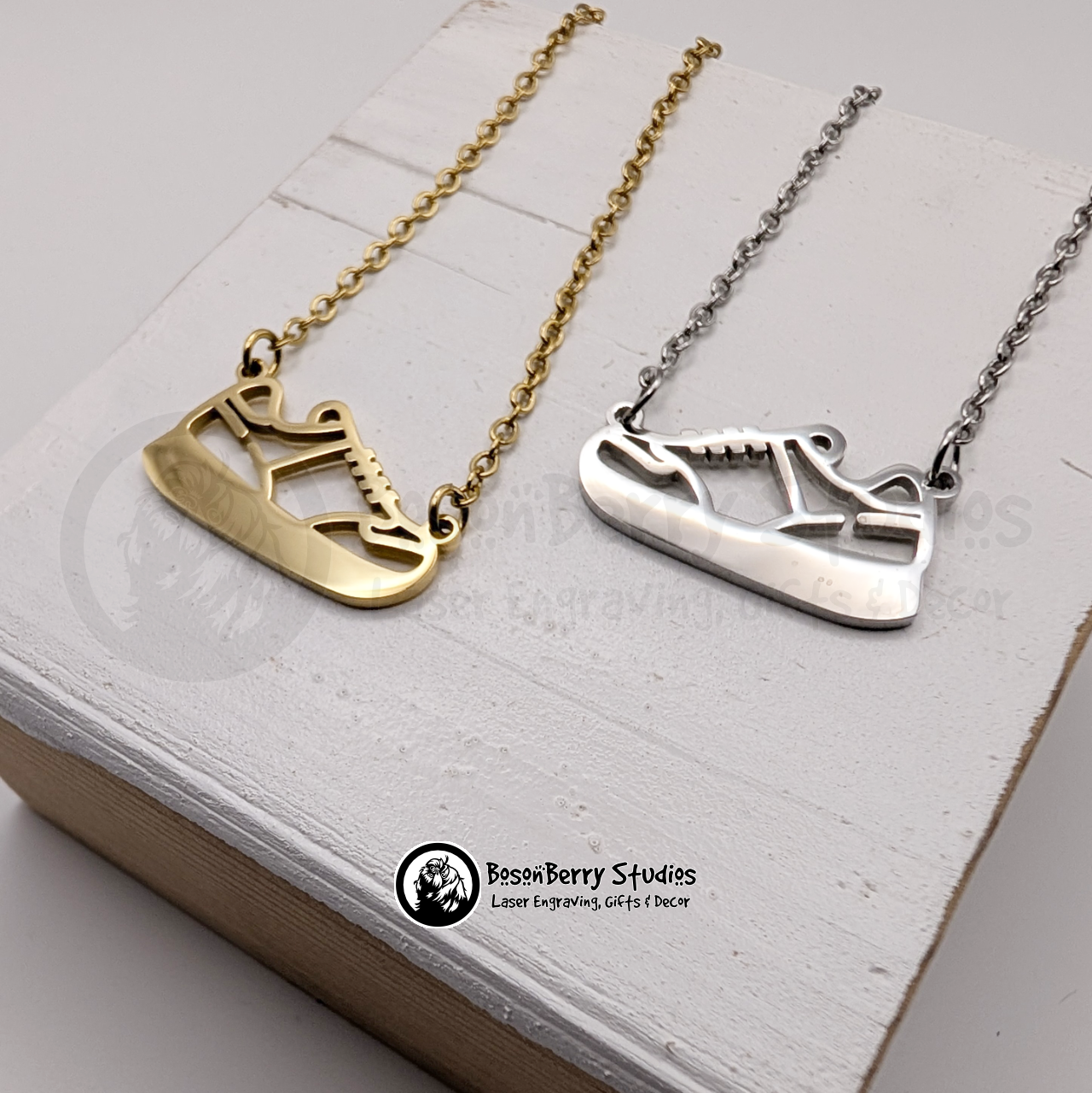 Two necklaces, one gold and one silver, with pendants shaped like sneakers on a white wooden stand with 'BosonBerry Studios' branding.