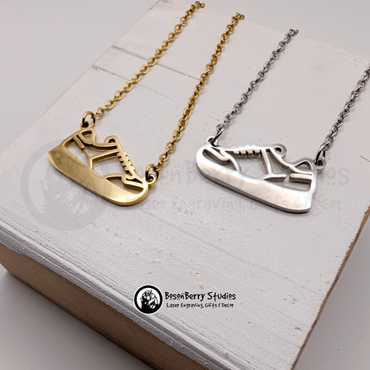 Two necklaces, one gold and one silver, with pendants shaped like sneakers on a white wooden stand with 'BosonBerry Studios' branding.