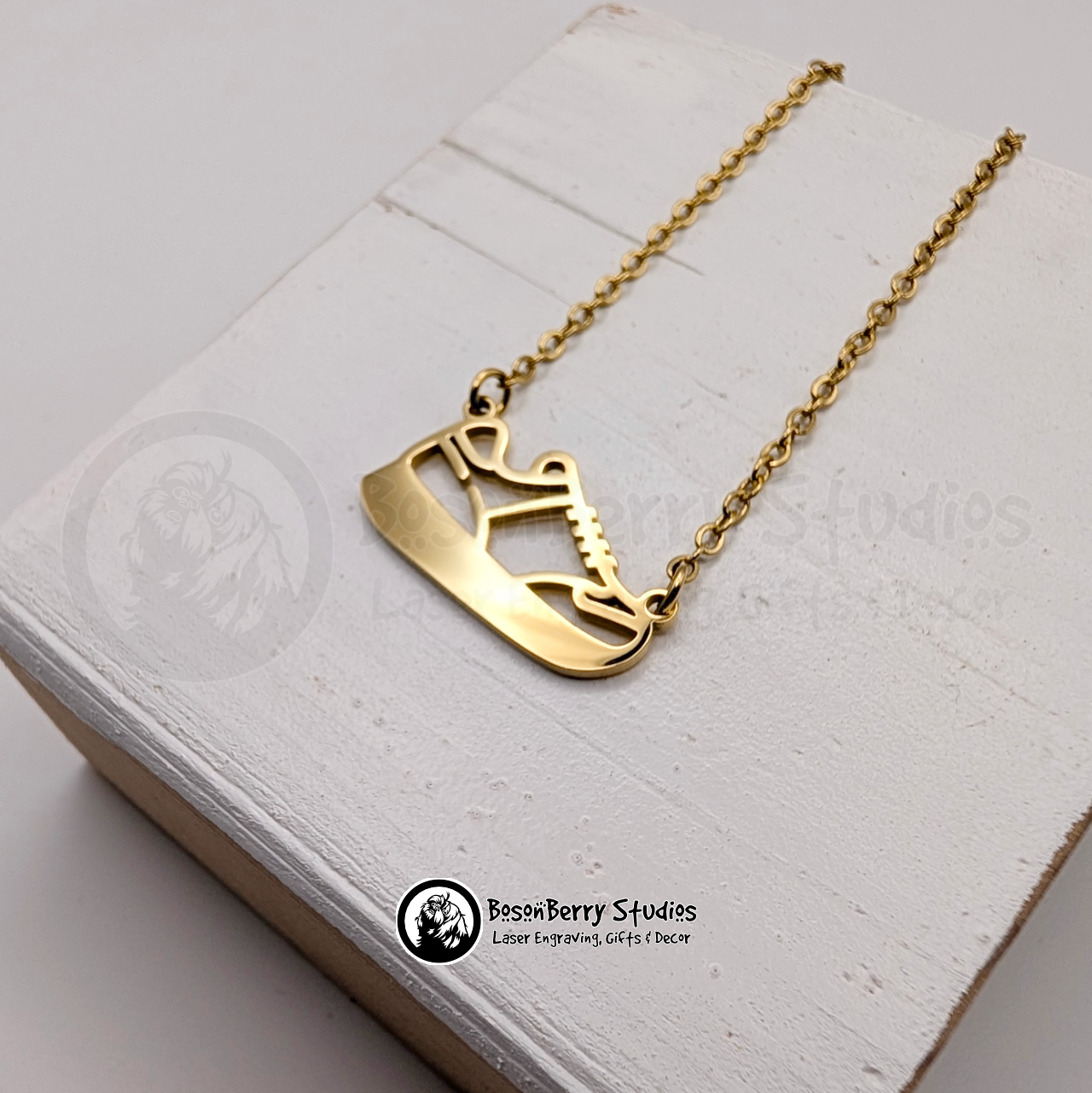 Gold necklace with a running shoe pendant on a white wooden stand with 'BosonBerry Studios' branding.