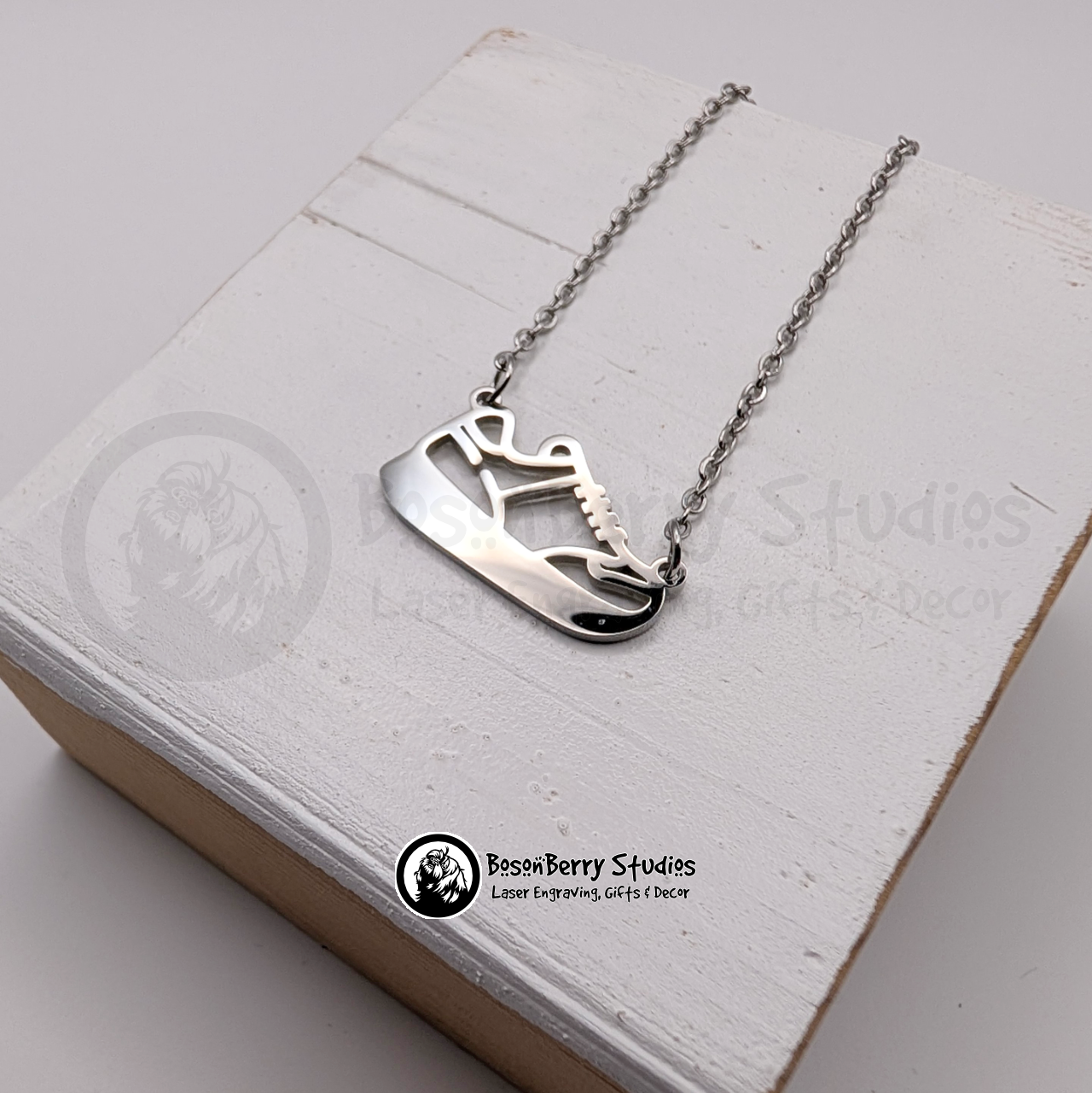 Silver necklace with a running shoe pendant on a white wooden stand with 'BosonBerry Studios' branding.
