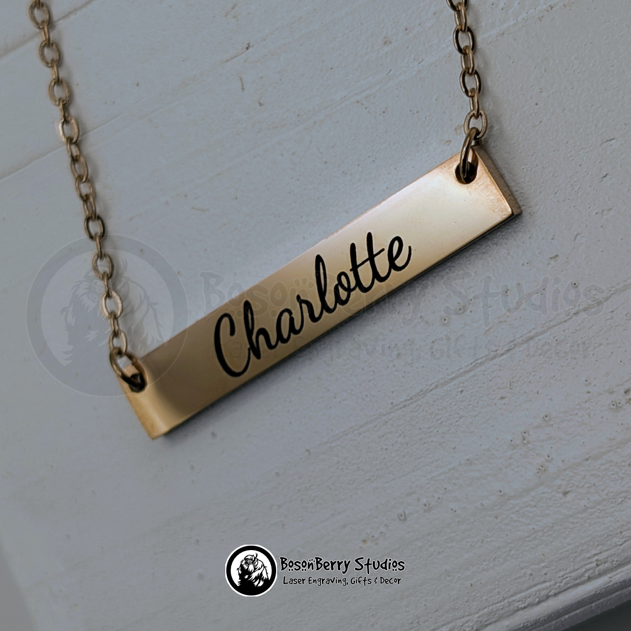 Gold bar necklace with 'Charlotte' engraving in black cursive font, on a white wooden stand with 'BosonBerry Studios' branding.