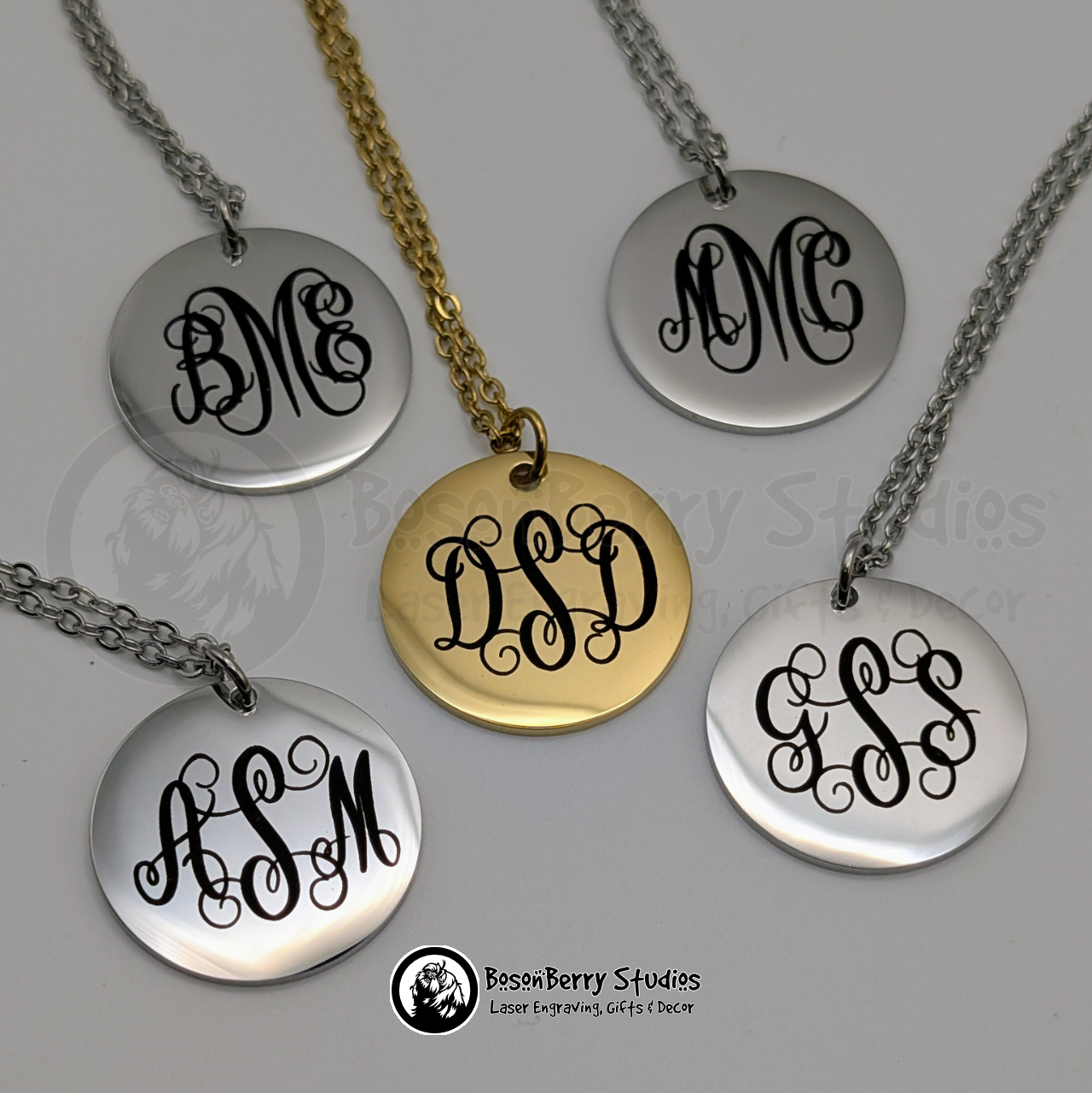 Five monogrammed round pendant necklaces, one gold and four silver, engraved with black curly font with different letter combinations, on a white background with 'BosonBerry Studios' branding.