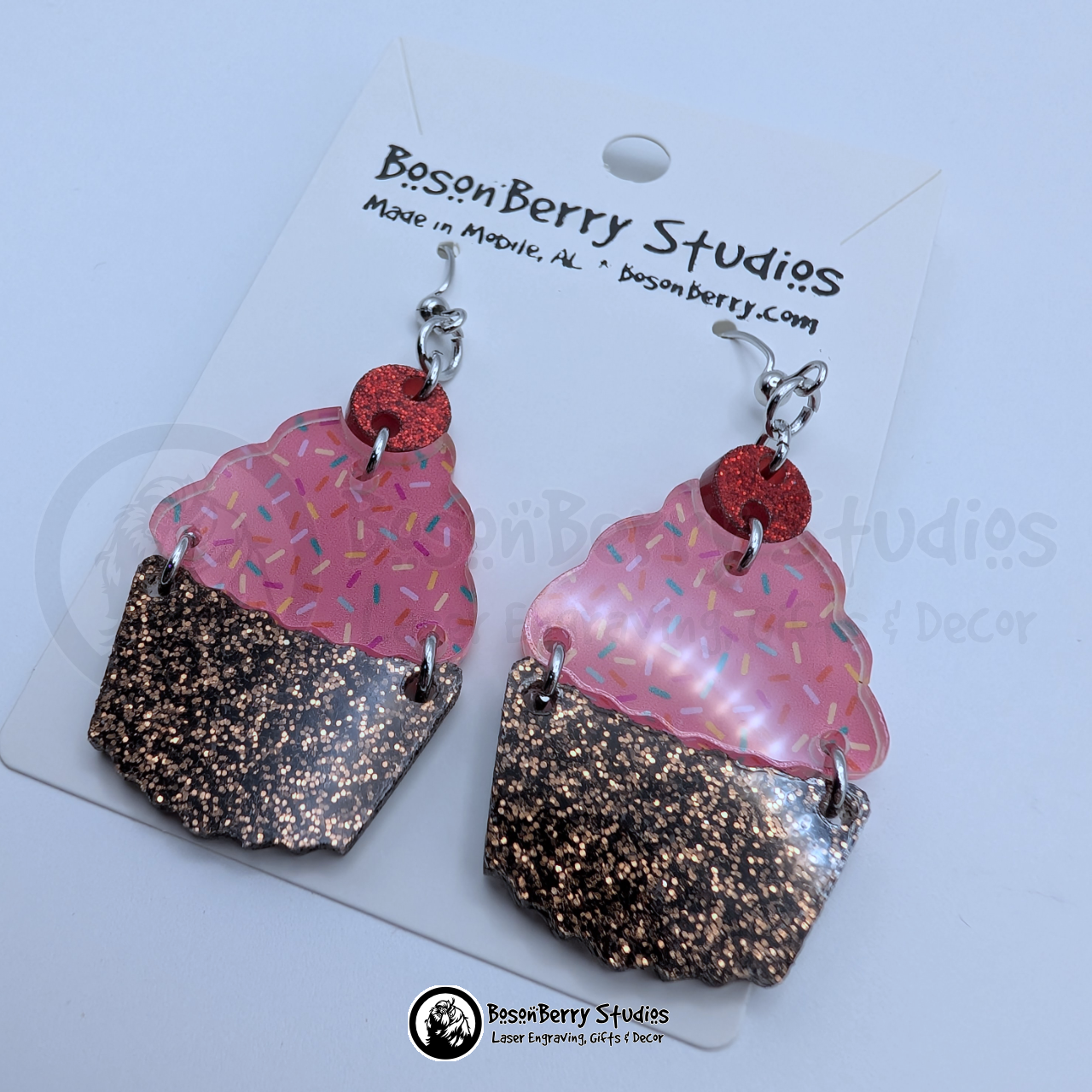 Tiered acrylic earrings with a glitter chocolate base, a pink with colored sprinkles icing center, and a red glitter cherry top, on a BosonBerry Studios branded display card.
