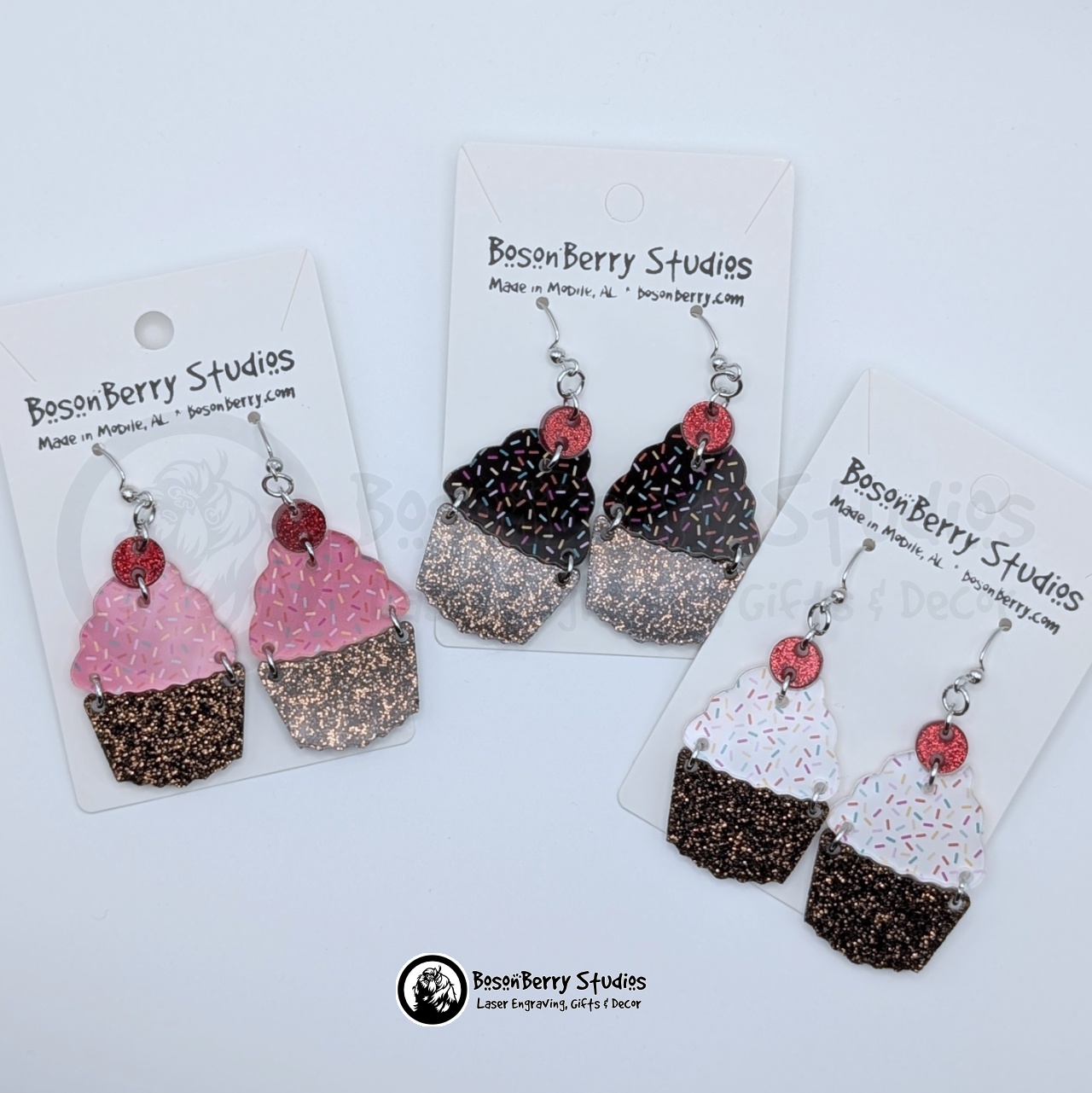 Trio of tiered acrylic earrings with a glitter chocolate base, a colored sprinkles icing center, and a red glitter cherry top, on BosonBerry Studios branded display cards.