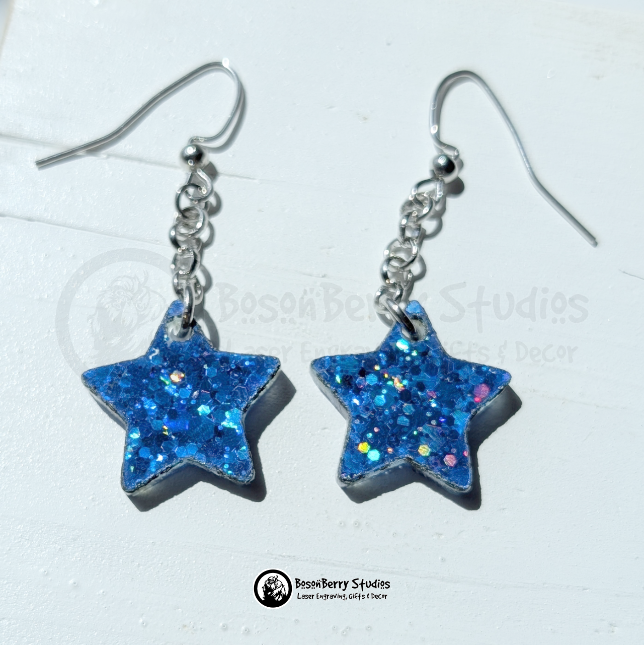 Blue star-shaped glitter dangle earrings on a white background with "BosonBerry Studios" branding.